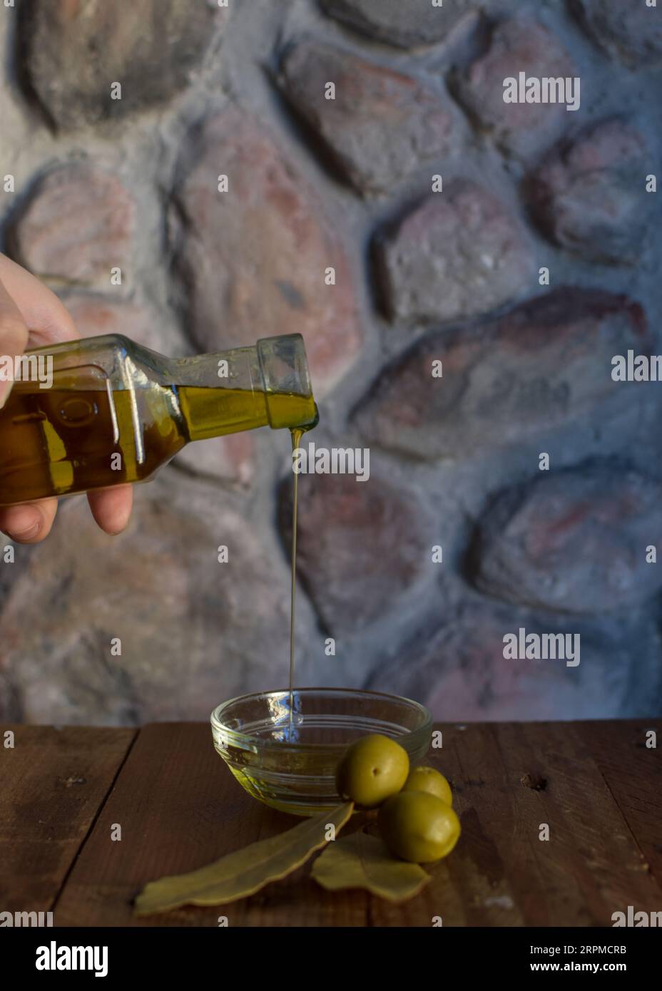 Eine Glasflasche Olivenöl steht auf einem Holztisch mit Oliven und einer strukturierten Steinmauer im Hintergrund, die ein rustikales Ambiente schafft. Stockfoto