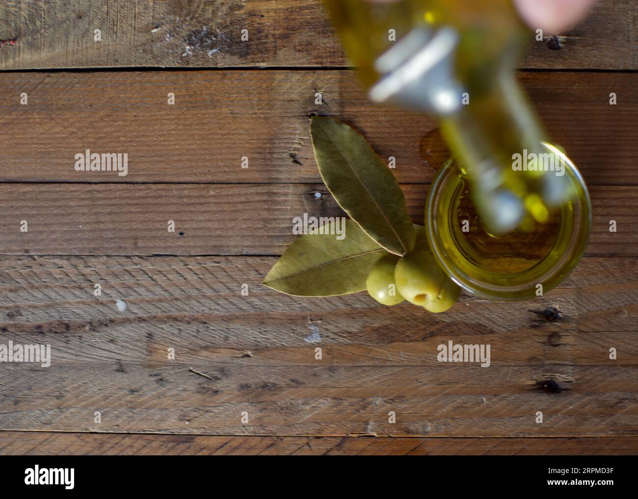 Eine Glasflasche Olivenöl steht auf einem Holztisch mit Oliven und einer strukturierten Steinmauer im Hintergrund, die ein rustikales Ambiente schafft. Stockfoto