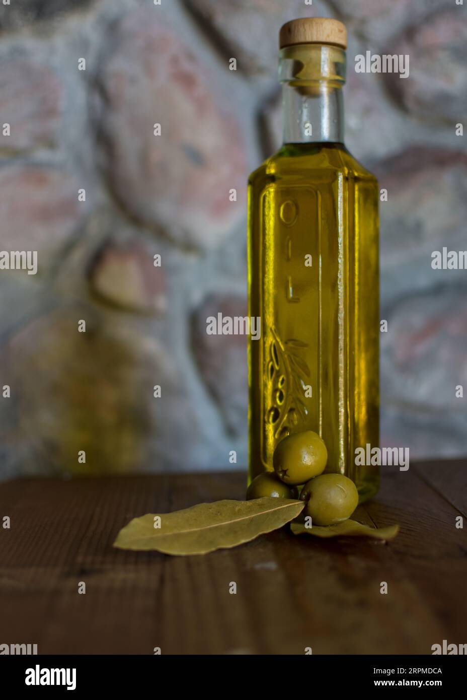 Eine Glasflasche Olivenöl steht auf einem Holztisch mit Oliven und einer strukturierten Steinmauer im Hintergrund, die ein rustikales Ambiente schafft. Stockfoto
