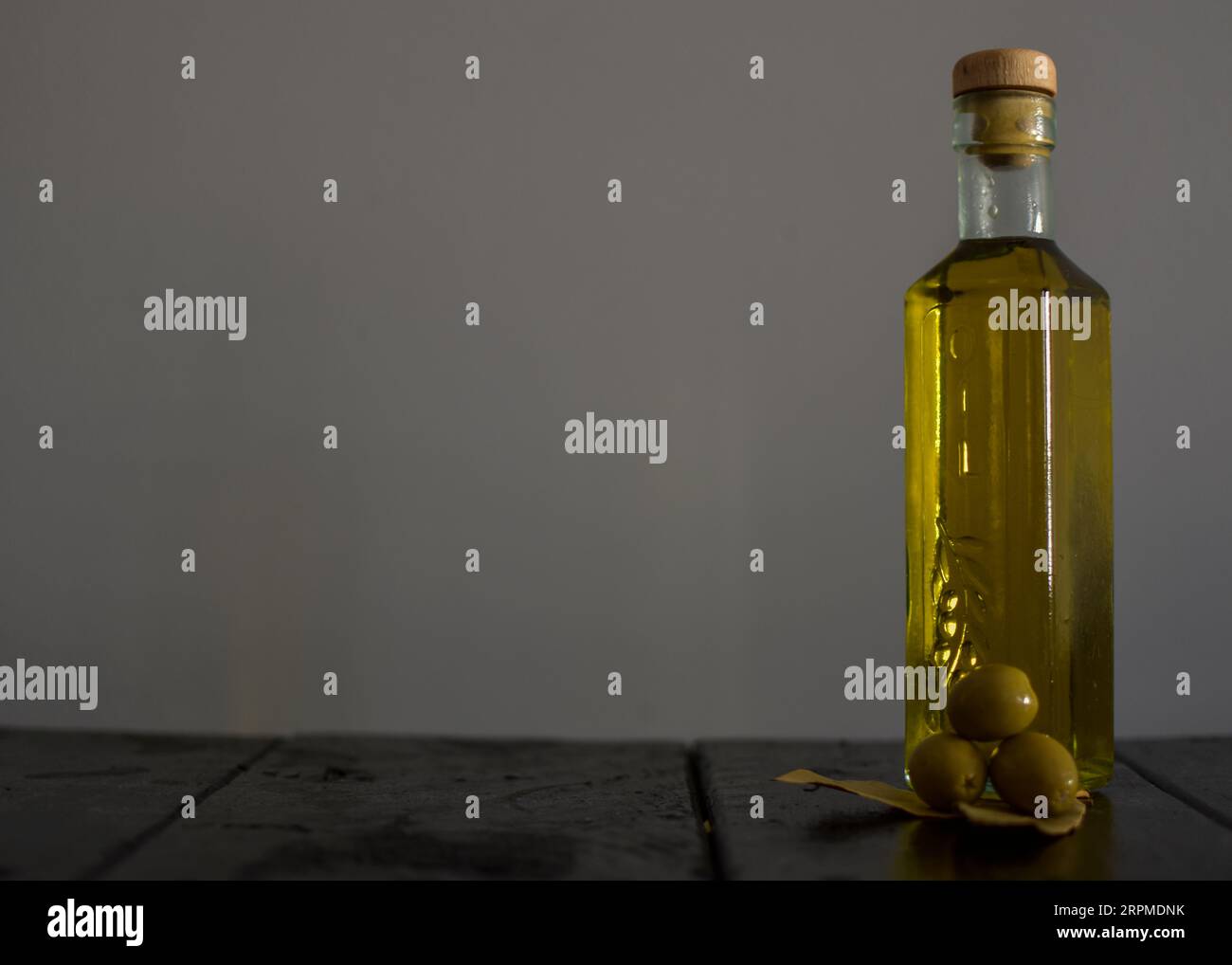 Eine Glasflasche Olivenöl steht auf einem Holztisch mit Oliven und einer strukturierten Steinmauer im Hintergrund, die ein rustikales Ambiente schafft. Stockfoto