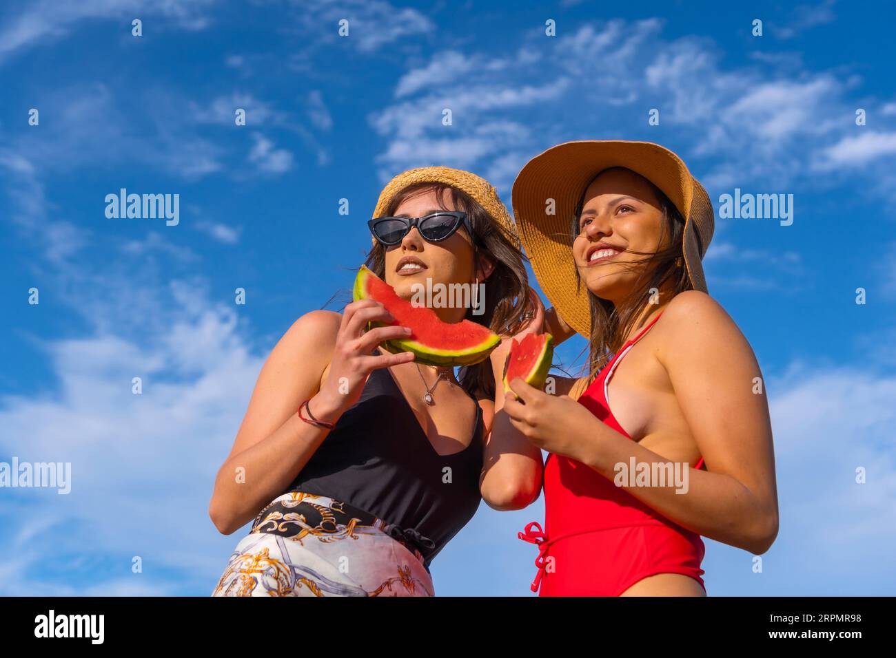 Porträt weiblicher Freunde am Strand im Sommer lächelnd beim Essen einer Wassermelone mit dem Himmel im Hintergrund, Bottom-up-Aufnahme Stockfoto