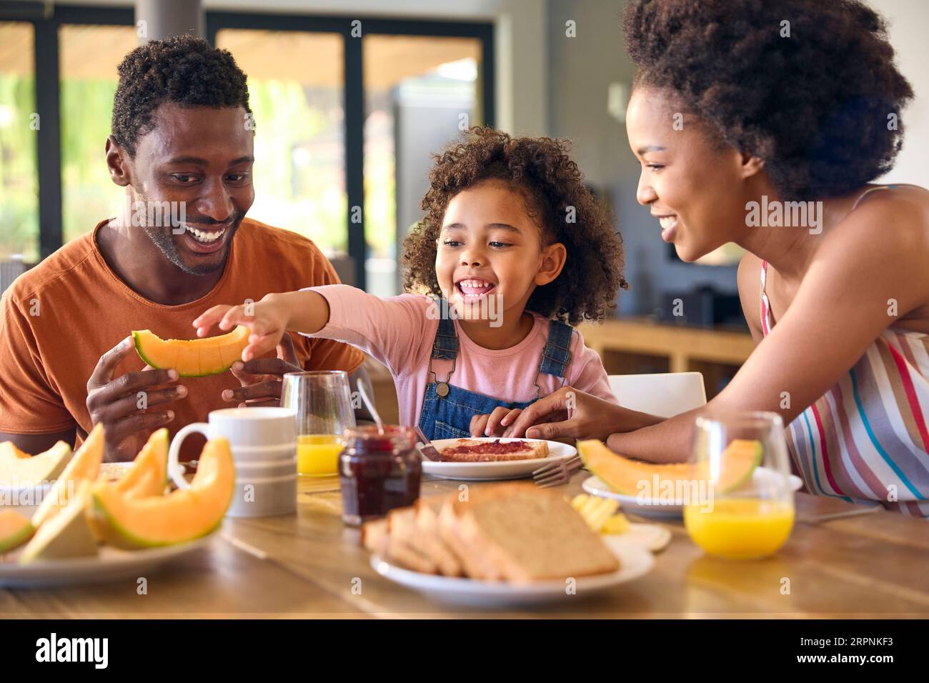 Familienschoß Mit Eltern Und Tochter Zu Hause Beim Frühstück, Das Marmelade Auf Brot Am Tisch Verteilt Stockfoto