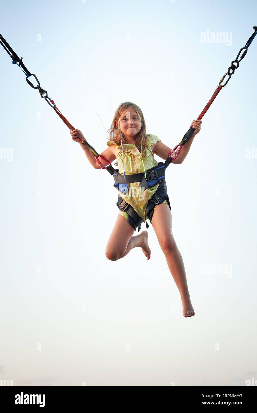 Bungee-Jumping auf Trampolin. Kleines Mädchen hüpft auf Bungee-Jumping im Vergnügungspark auf Sommerferien. Kind fliegt über den blauen Himmel, während es weiterspringt Stockfoto