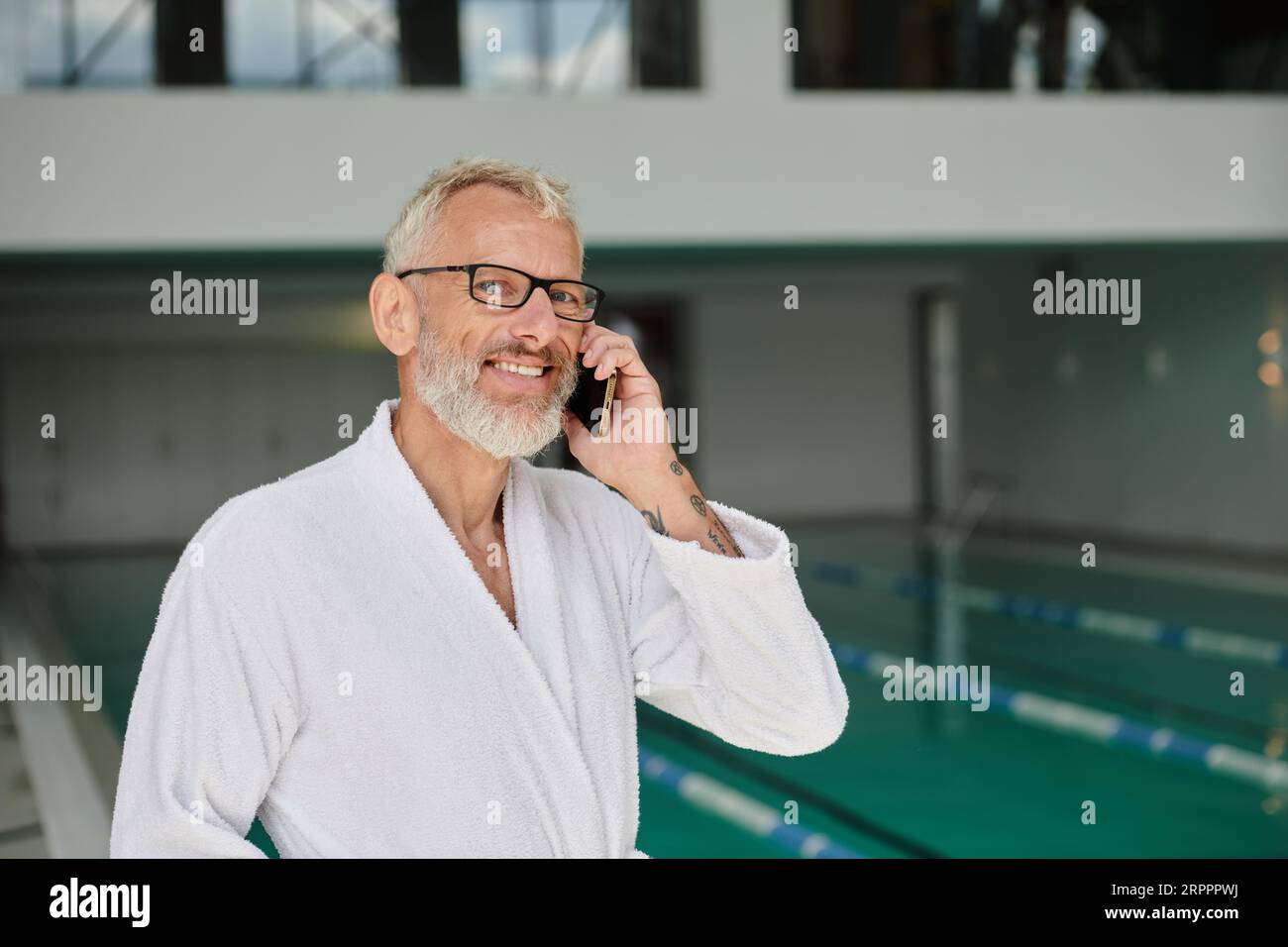 Positiver reifer Mann in weißem Gewand und Brille, der auf dem Smartphone im Spa-Center spricht, Retreat Stockfoto