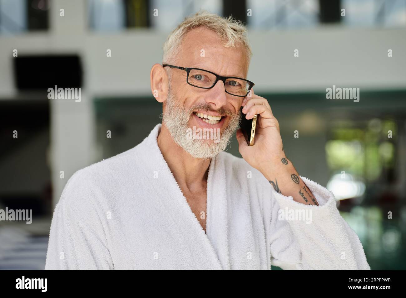 Positiver reifer Mann in weißem Bademantel und Brille, der auf dem Smartphone im Spa-Center spricht Stockfoto
