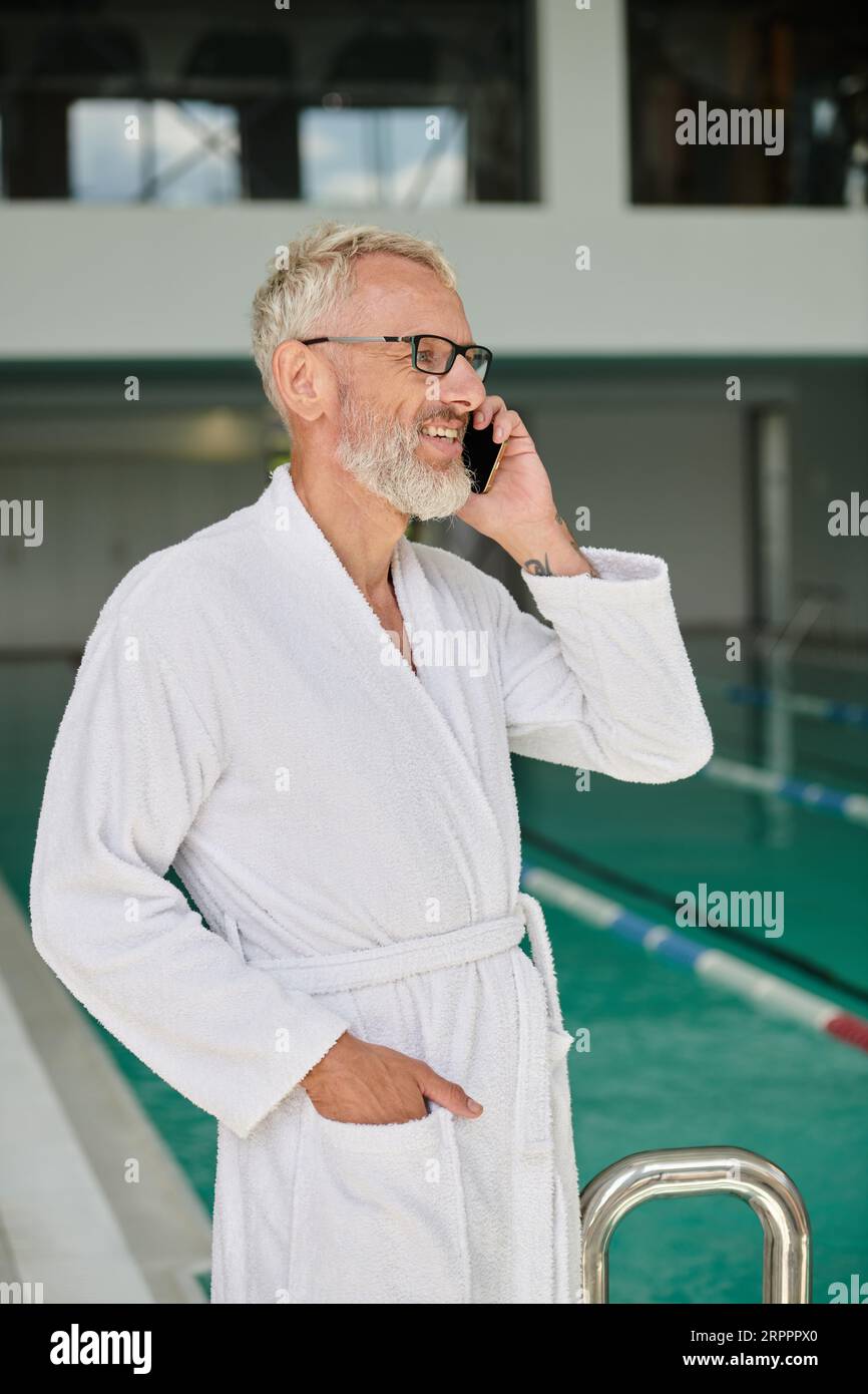 Fröhlicher reifer Mann in weißem Bademantel und Brille, der auf dem Smartphone im Spa-Center spricht, Retreat Stockfoto