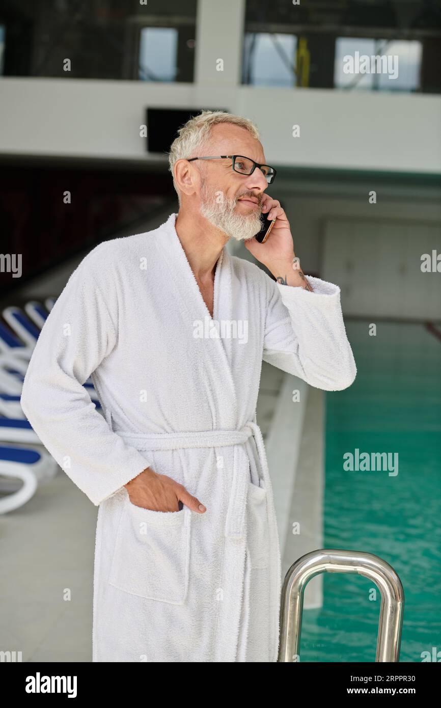 Tätowierter reifer Mann in weißem Bademantel und Brille, die auf dem Smartphone im Spa-Center sprechen, Retreat Stockfoto