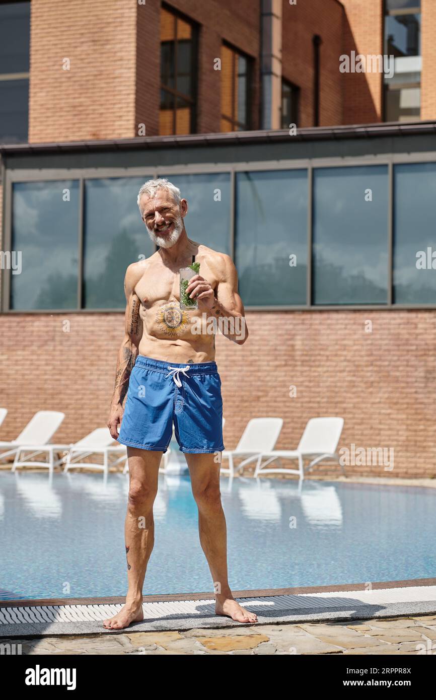 Tätowierter Mann mittleren Alters in kurzen Hosen, lächelnd und mit Mojito-Cocktail am Pool, Wellness-Retreat Stockfoto