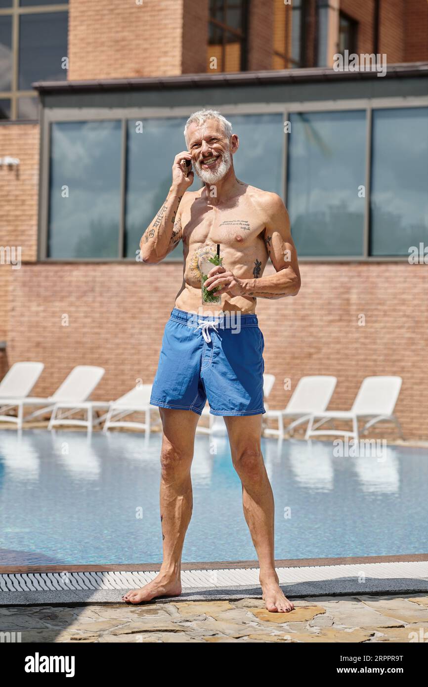 Glücklicher reifer Mann in Shorts, der auf dem Smartphone spricht und einen Cocktail am Pool hält, Wellness-Retreat Stockfoto