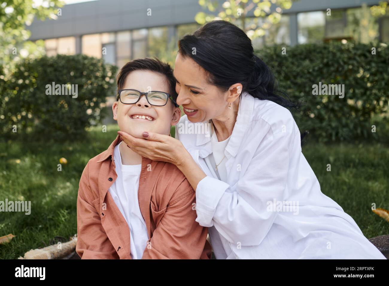Glückliche Frau mittleren Alters, die das Gesicht des Sohnes mit Down-Syndrom im Park berührt, bedingungslose Liebe Stockfoto
