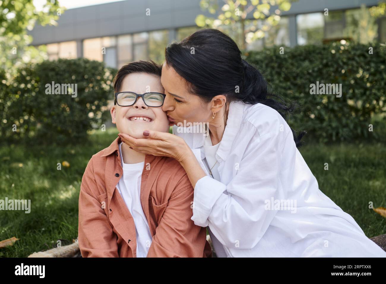 Frau mittleren Alters, die einen sorglosen Sohn mit Down-Syndrom küsst und mit geschlossenen Augen im Park lächelt Stockfoto