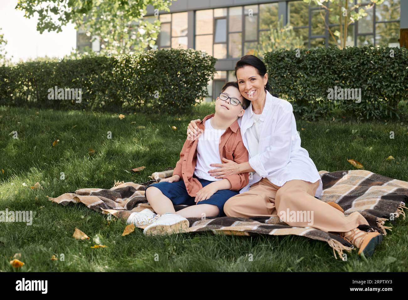 Eine lächelnde Frau mittleren Alters, die einen jungen Sohn mit Down-Syndrom auf einer Decke im Park umarmt, gute Zeit Stockfoto