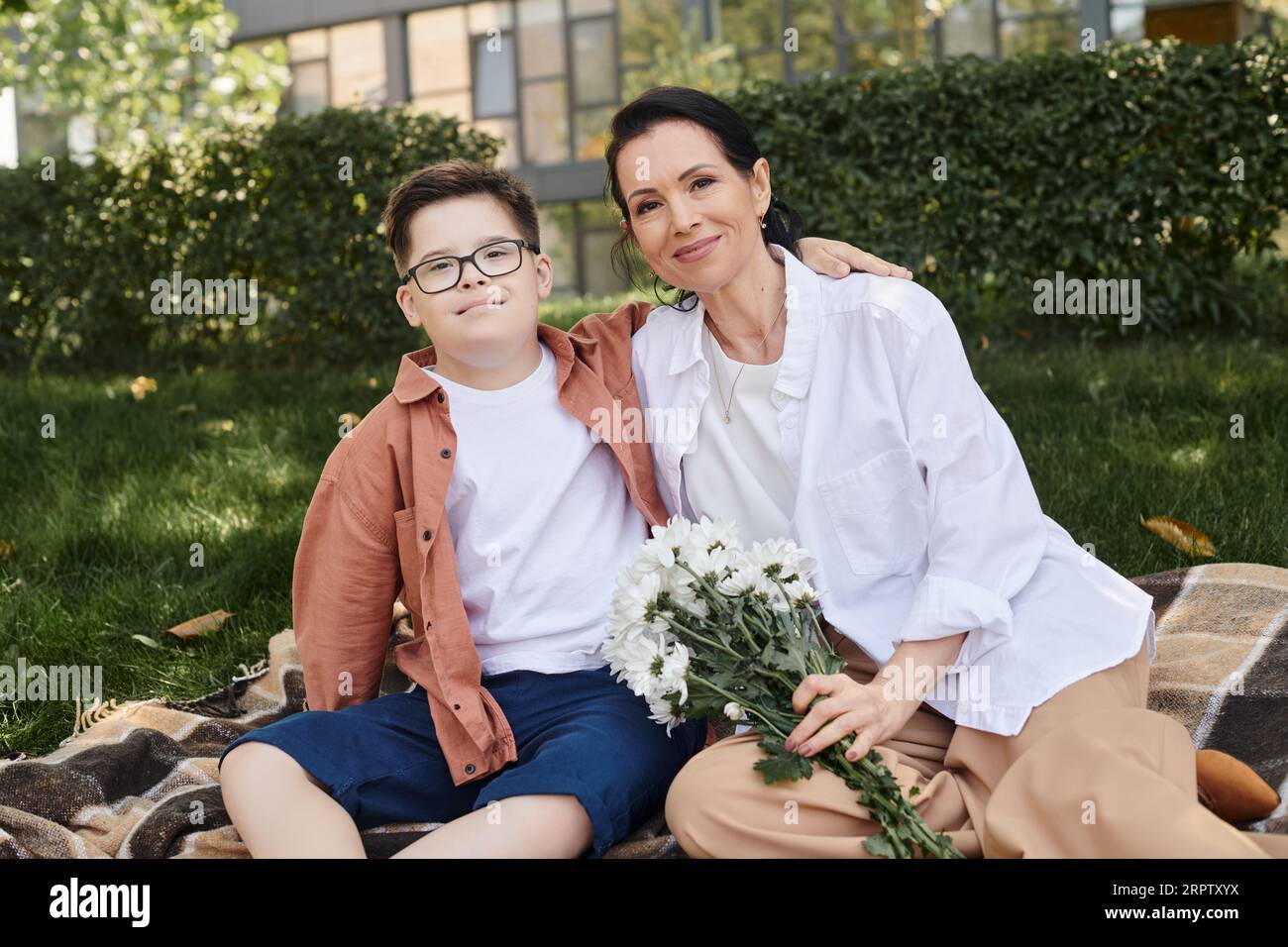 Frau mittleren Alters, die Blumen hält, während sie in der Nähe des Sohnes sitzt, mit Down-Syndrom auf einer Decke im Park Stockfoto