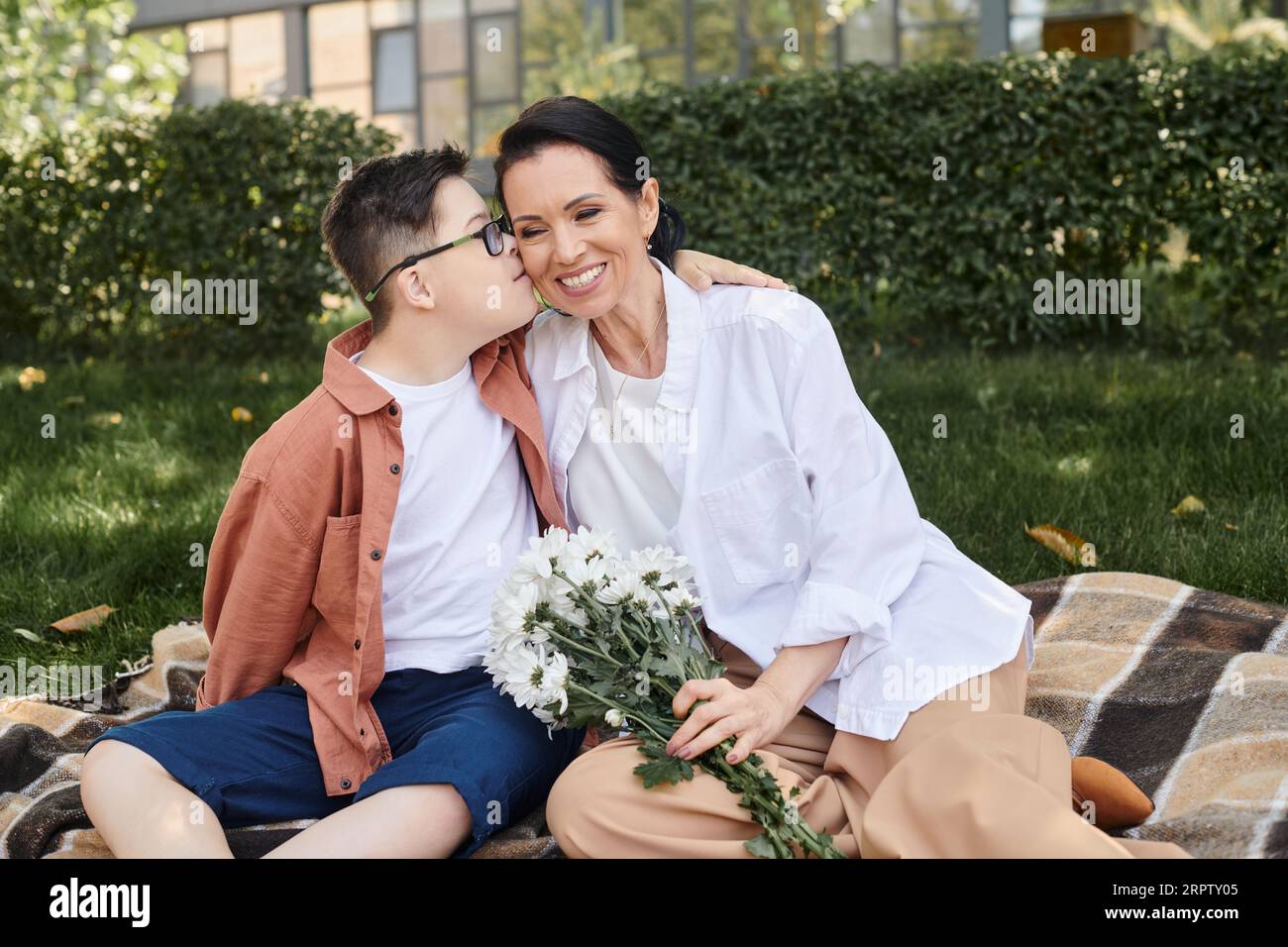 Kind mit Down-Syndrom, das Mutter küsst, die mit Blumen auf einer Decke im Park sitzt, bedingungslose Liebe Stockfoto