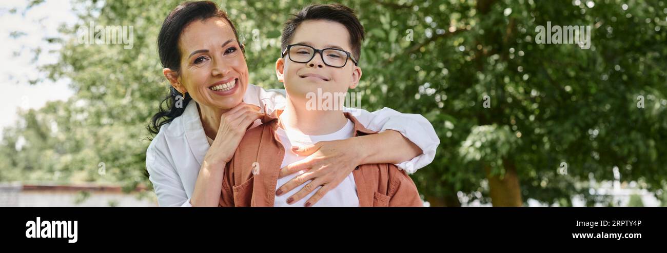 Lächelnde Frau mittleren Alters umarmt Sohn mit Down-Syndrom im Park, emotionale Verbindung, Banner Stockfoto