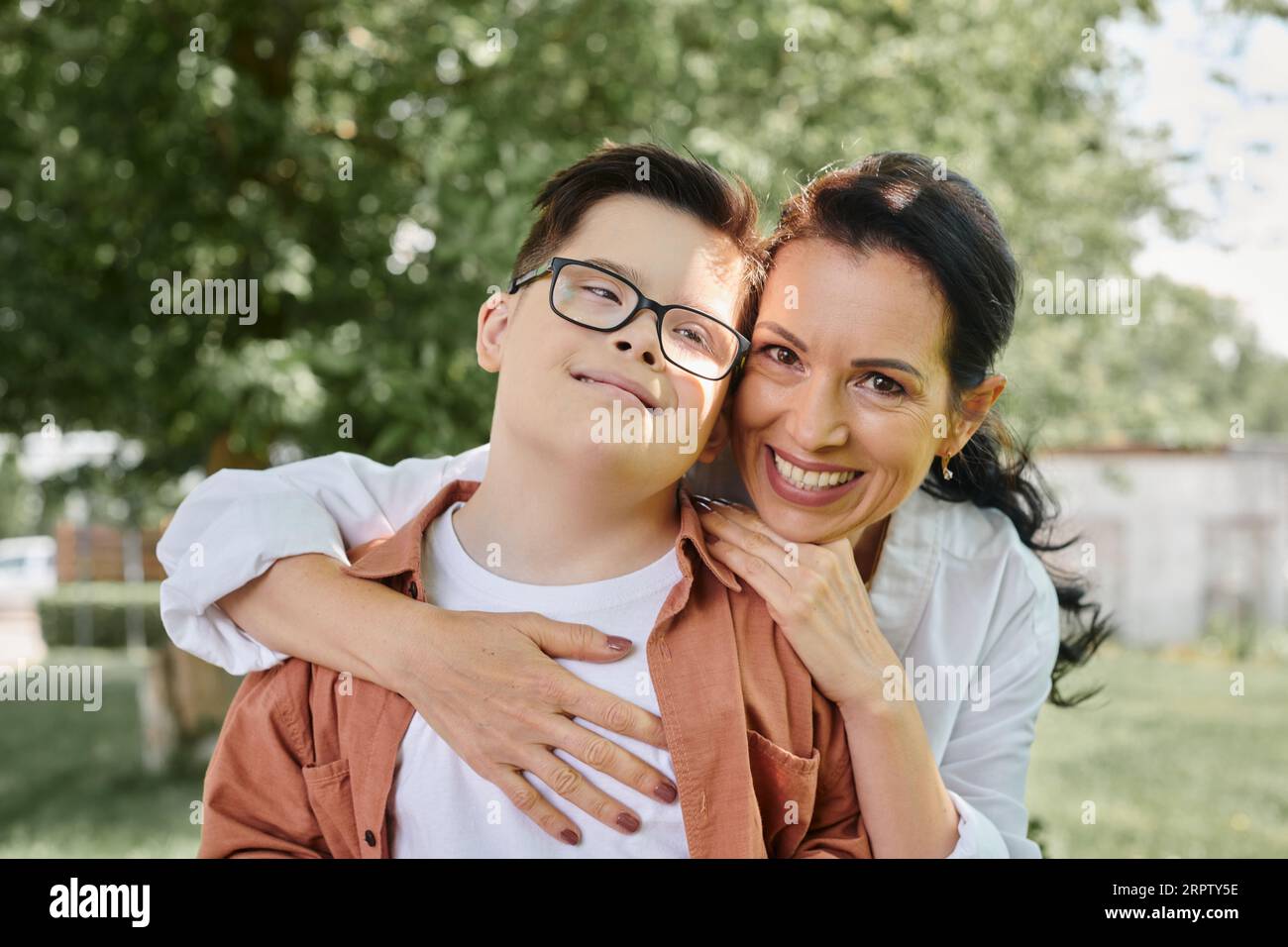 Glückliche Frau mittleren Alters, die einen Jungen mit Down-Syndrom umarmt und im Park lächelt, besondere Familie Stockfoto
