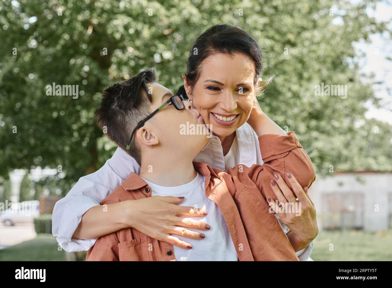 Kind mit Down-Syndrom, das eine lächelnde Mutter mittleren Alters während eines Parkausflugs küsst, glückliche Momente Stockfoto