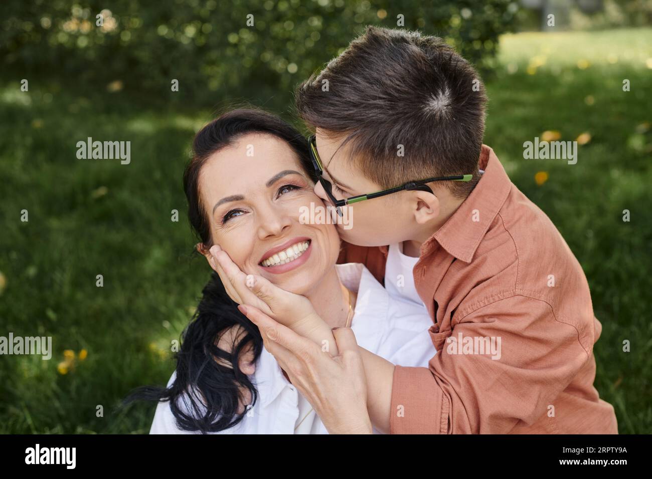 Junge mit Down-Syndrom, in Brille, küssende überfreuliche Mutter im Park, bedingungslose Liebe Stockfoto