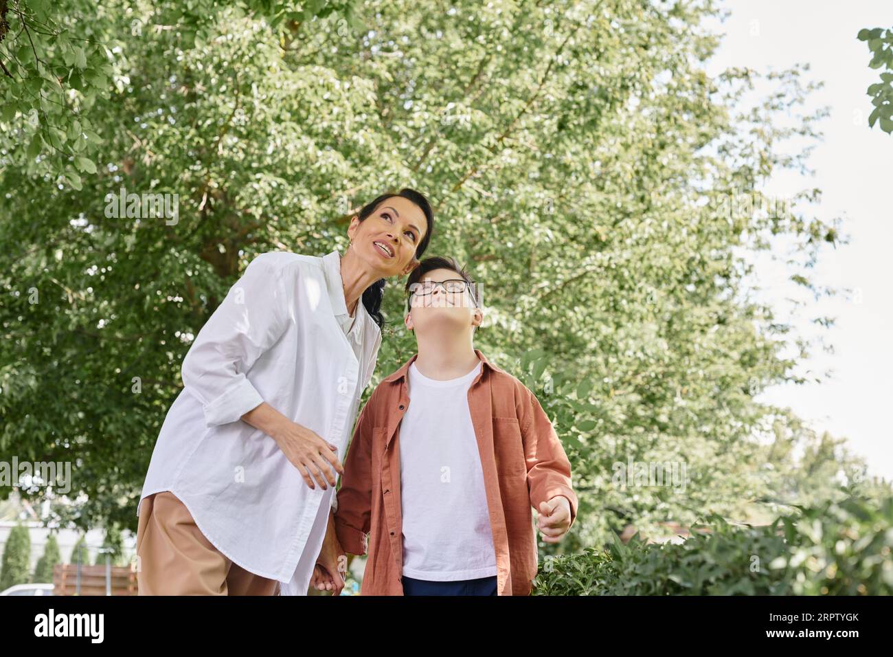 Lächelnde Frau mittleren Alters und Junge mit Down-Syndrom, die im Park wegschauen, glückliche Momente Stockfoto