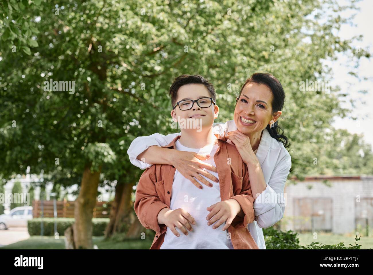 Fröhliche Frau mittleren Alters, die lächelnden Sohn mit Down-Syndrom im Park umarmt, bedingungslose Liebe Stockfoto