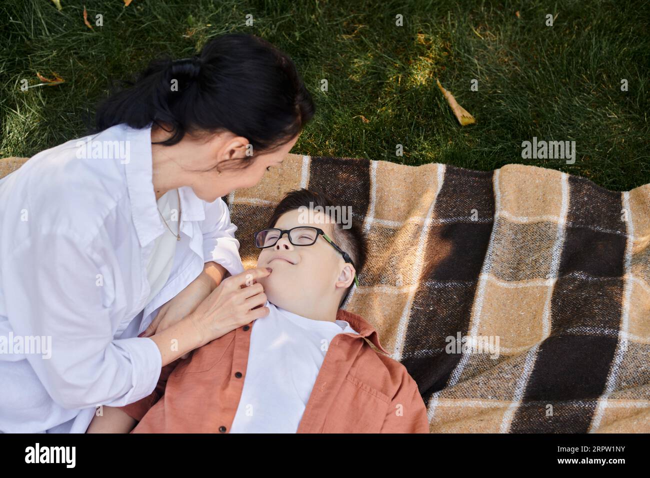 Draufsicht der Frau, die das Gesicht des Sohnes mit Down-Syndrom auf der Decke im Park berührt, Liebe und Pflege Stockfoto