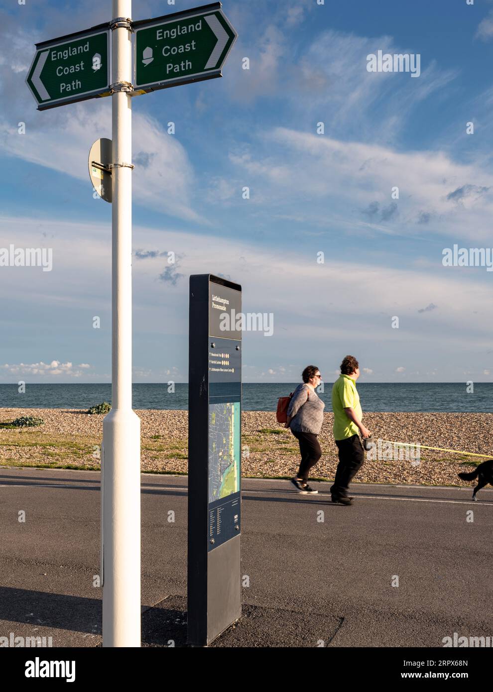 Promenade in Littlehampton, West Sussex, Großbritannien; zwei Personen gehen zu Fuß auf das Meer und das Küstenpfadschild im Vordergrund Stockfoto