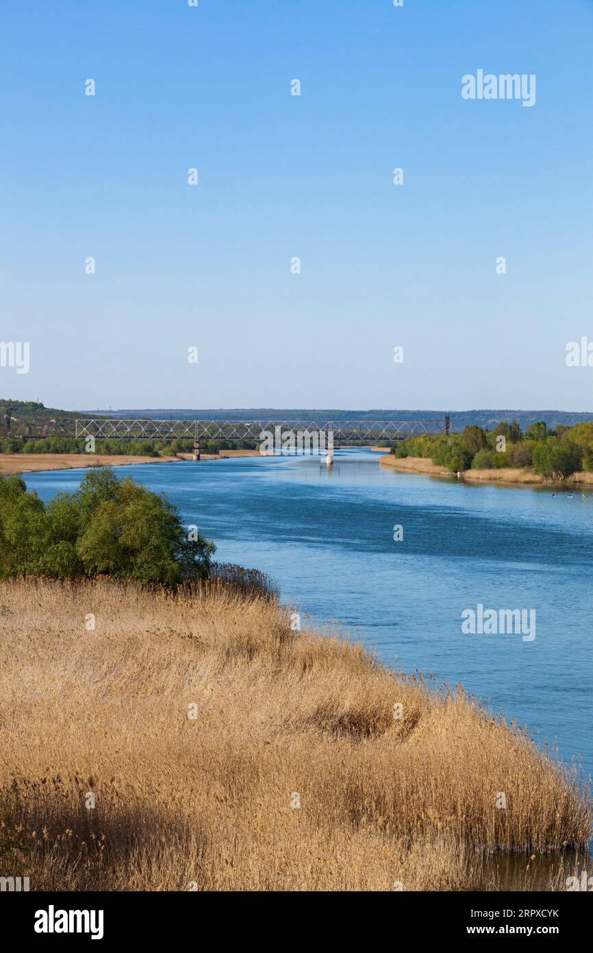 Dniester. Blick auf den Fluss von der Eisenbahnbrücke. Der blaue Himmel spiegelt sich im Wasser. Grüne Bäume und trockenes Schilf. Vertikales Foto. Stockfoto