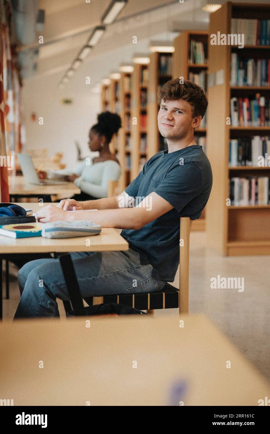 Porträt eines lächelnden Schülers, der während des Studiums in der Bibliothek am Tisch sitzt Stockfoto