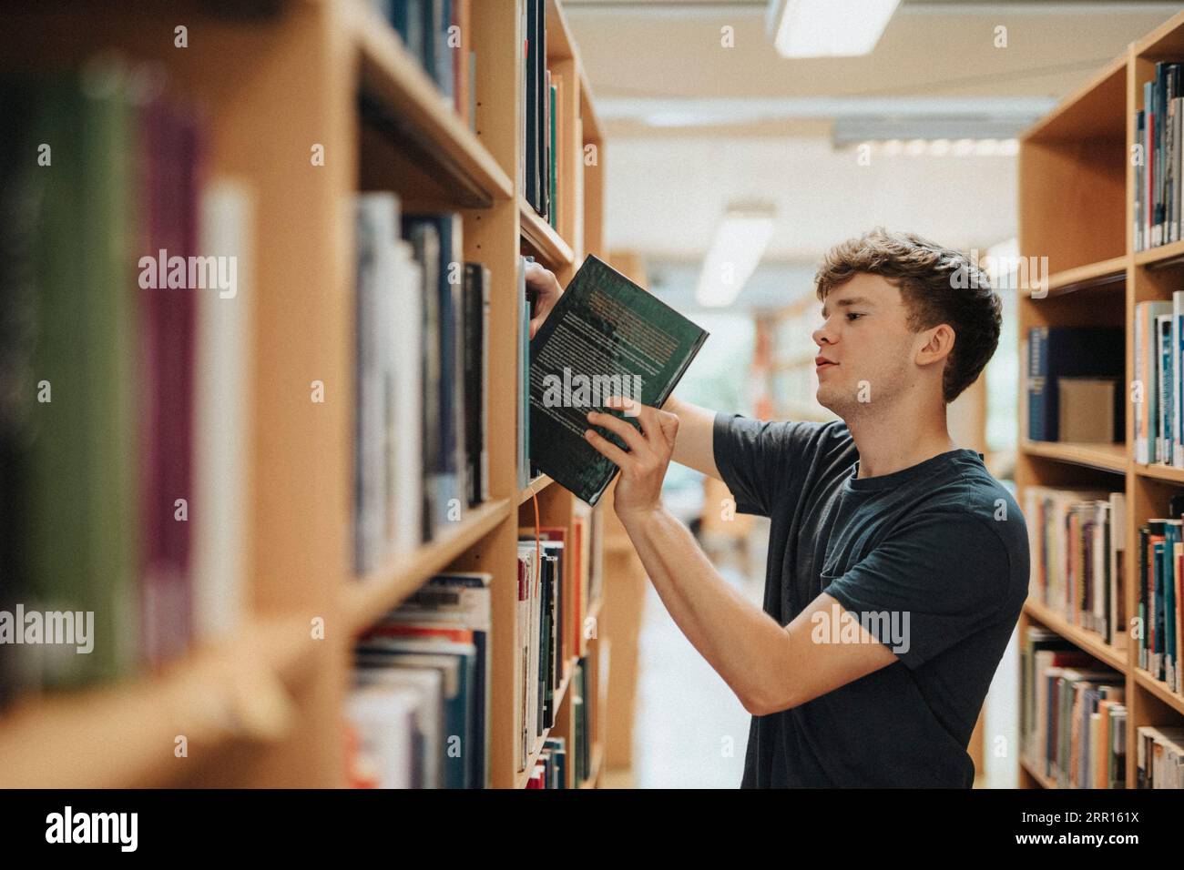 Seitenansicht eines männlichen Studenten, der das Buch aus dem Bücherregal in der Universitätsbibliothek entfernt Stockfoto
