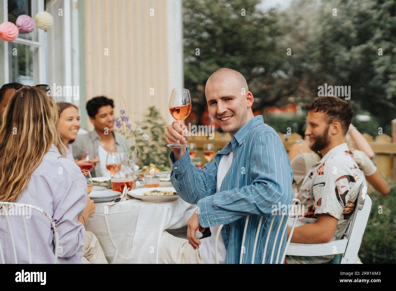Seitenansicht eines Porträts des lächelnden jungen Mannes, der Weinglas hält, während er mit Freunden auf dem Stuhl sitzt, während der Dinnerparty im Caf Stockfoto