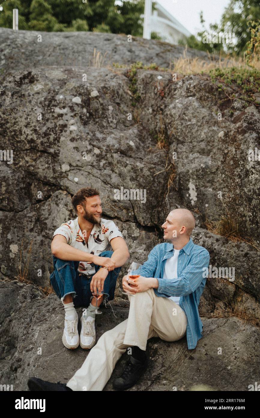 Junge männliche Freunde trinken Wein und reden miteinander, während sie auf Felsen sitzen Stockfoto