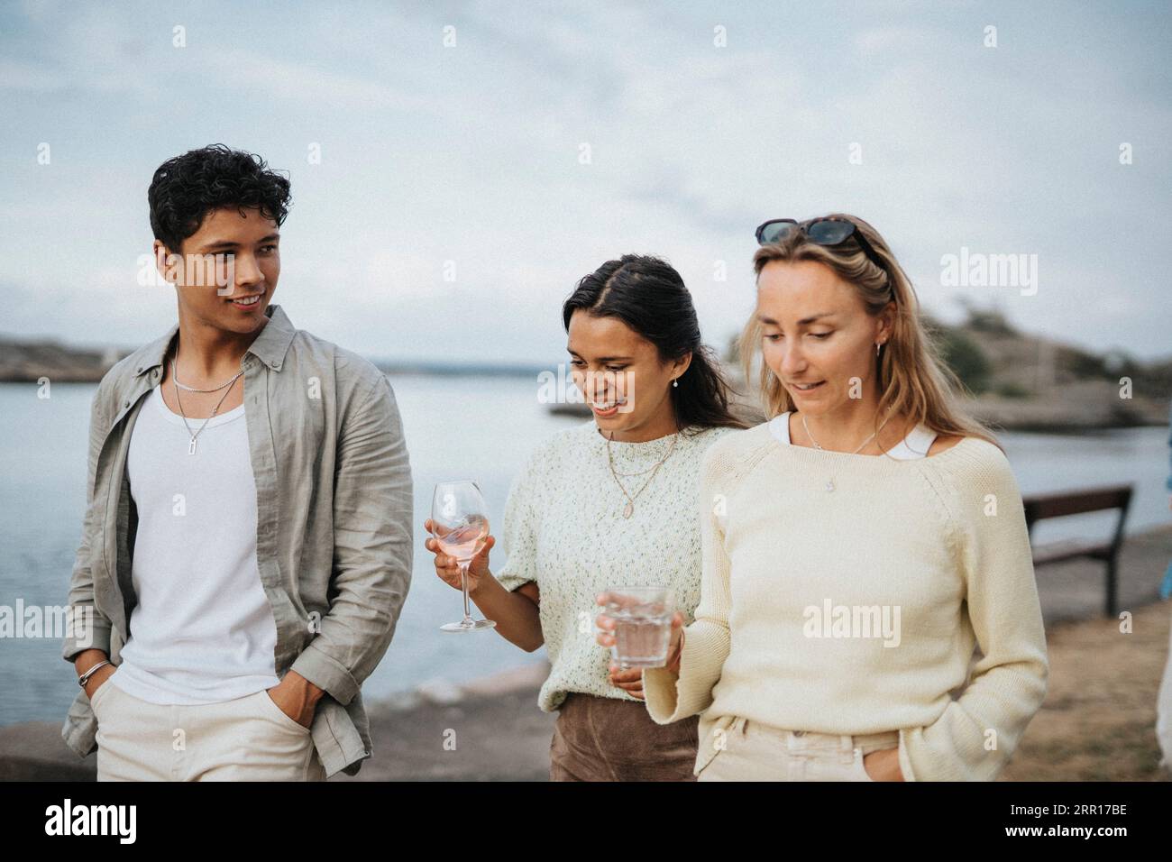 Mann, der mit Freundinnen spricht und Getränke hält, während er in der Nähe des Sees spaziert Stockfoto