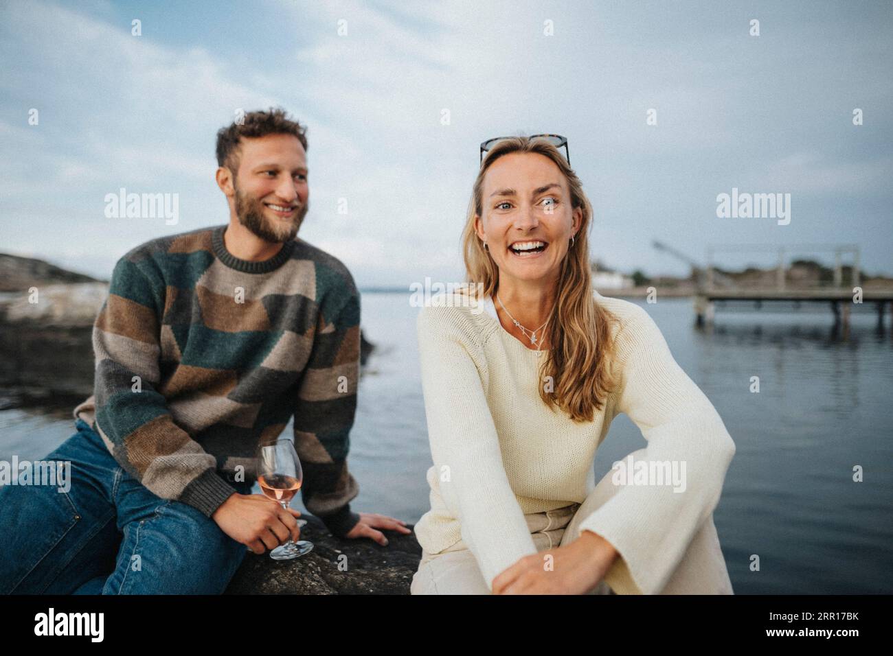 Glückliche Frau, die mit einem männlichen Freund sitzt und Wein trinkt, während sie auf einem Felsen in der Nähe des Sees sitzt Stockfoto