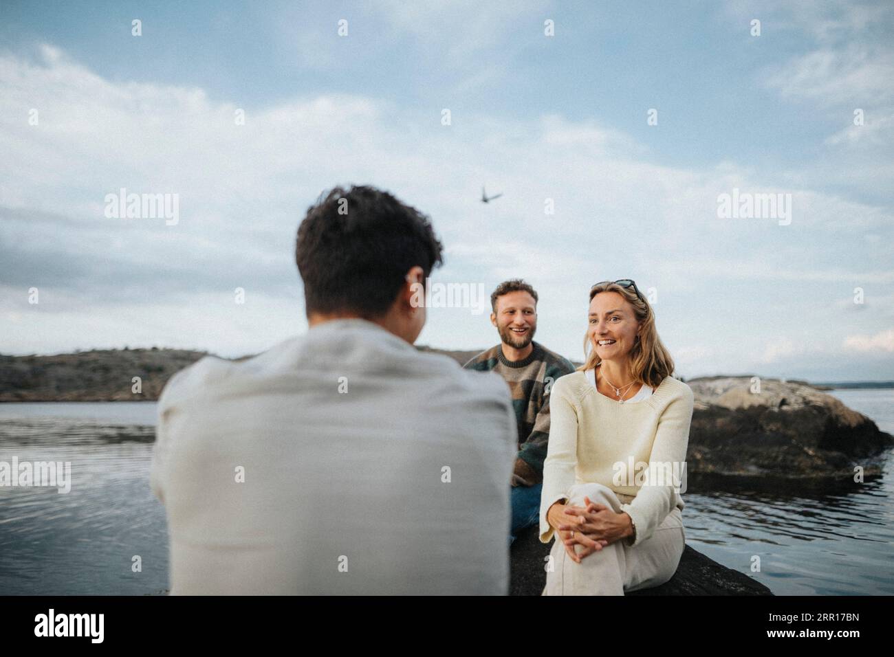 Lächelnde männliche und weibliche Freunde, die miteinander reden, während sie in der Nähe des Sees sitzen Stockfoto