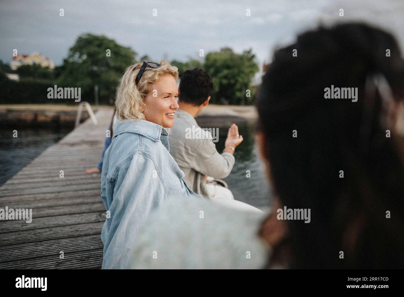 Lächelnde Frau, die wegschaut, während sie mit einem Freund auf dem Pier in der Nähe des Sees sitzt Stockfoto