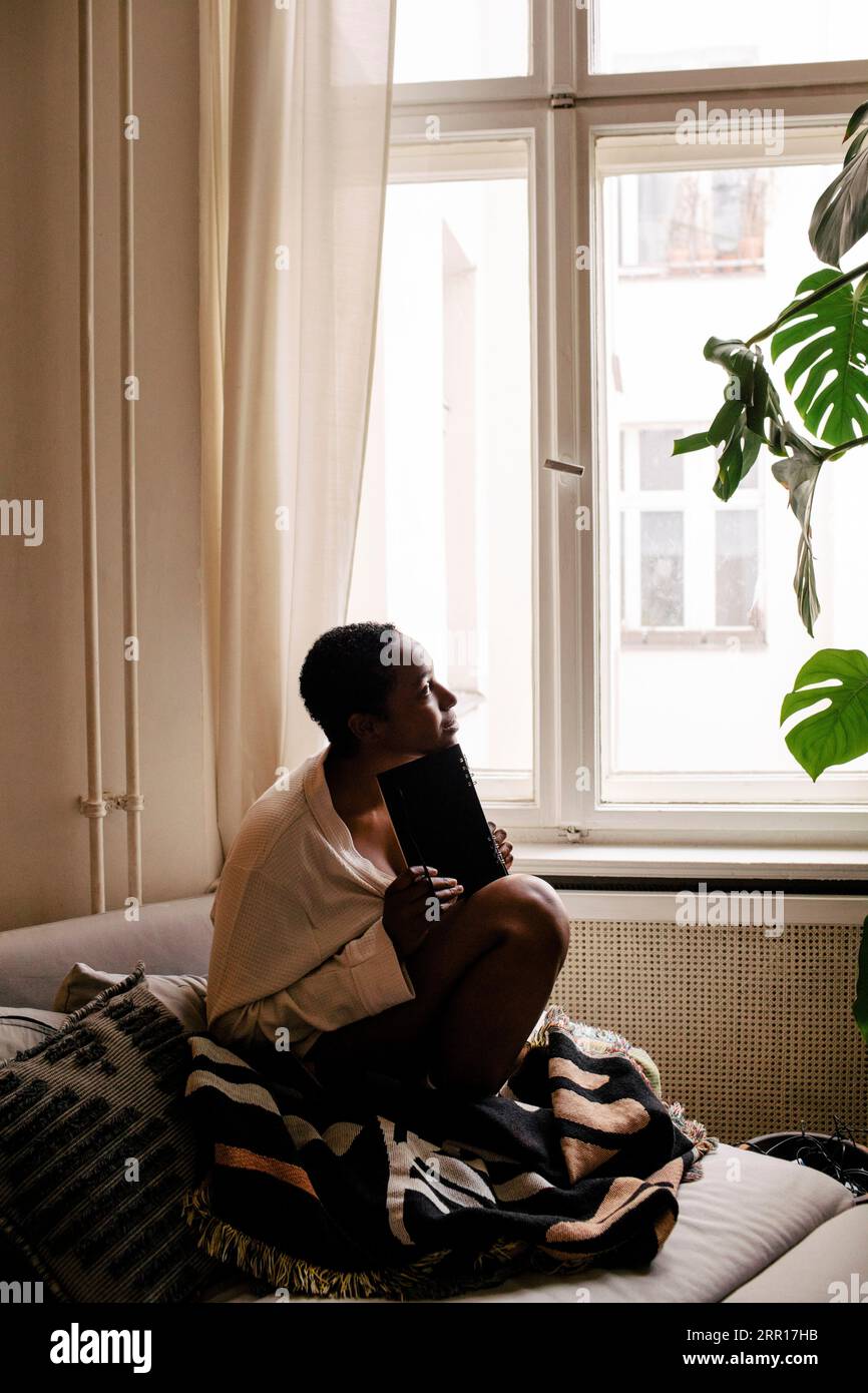 Nachdenkliche junge Frau mit Buch, das zu Hause auf dem Bett sitzt Stockfoto