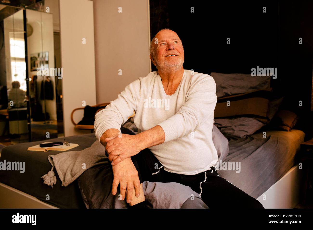 Lächelnder älterer Mann, der aufschaut, während er zu Hause auf dem Bett sitzt Stockfoto