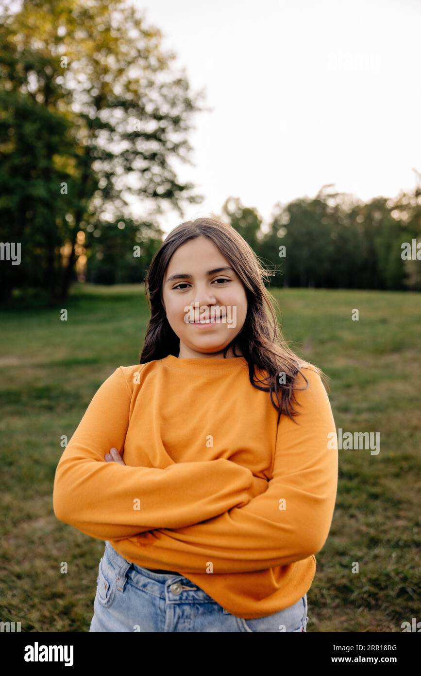 Porträt eines lächelnden Mädchens, das ein oranges T-Shirt trägt, das auf dem Spielplatz mit überkreuzten Armen steht Stockfoto