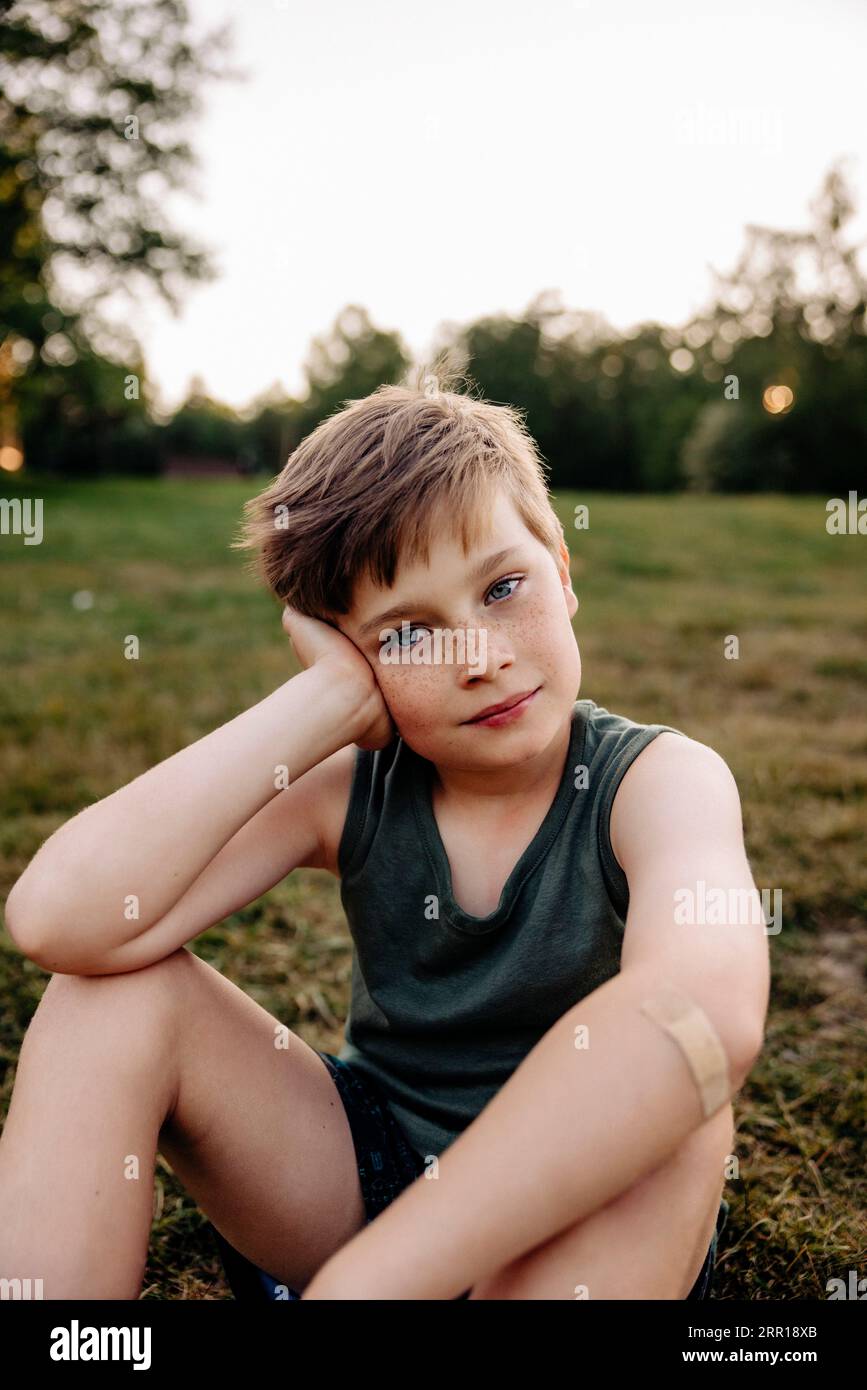 Porträt des Jungen, der sich auf dem Ellbogen lehnt, während er auf dem Rasen auf dem Spielplatz sitzt Stockfoto