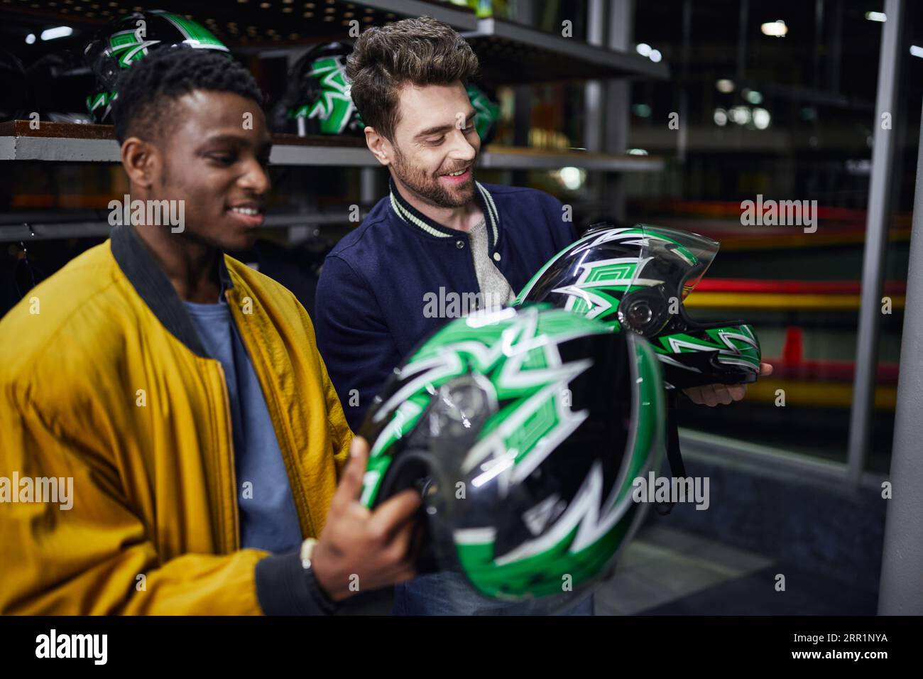 Zwei multikulturelle Freunde in Bomberjacken, die Helme im Inneren der Kartbahn betrachten, Go-Cart Stockfoto