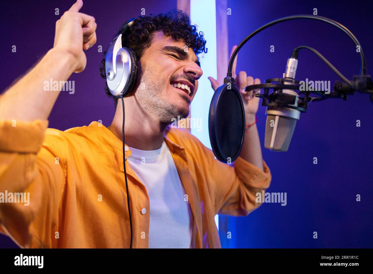 Seitenansicht eines männlichen professionellen Sängers in Kopfhörern, der in der Nähe des Mikrofons eine hohe Note nimmt und nach oben zeigt, während er im Studio Musik macht Stockfoto