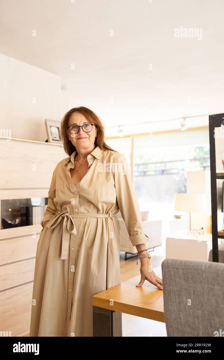 Positive reife Frau in trendigem Kleid und Brille lächelnd und in die Kamera blickend, während sie in der Nähe von Tisch und Regal im modernen Möbelgeschäft steht Stockfoto