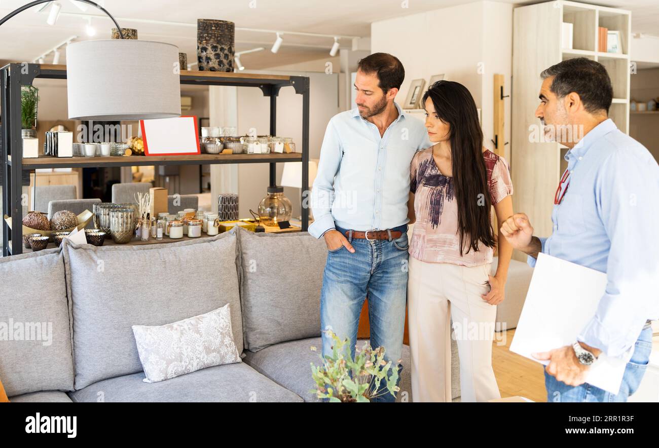 Konzentriertes junges Paar im legeren Kleidungsgeschäft im hellen Möbelgeschäft mit reifem männlichen Verkäufer in einem modernen Geschäft mit Sofa und Regalen bei Tageslicht Stockfoto