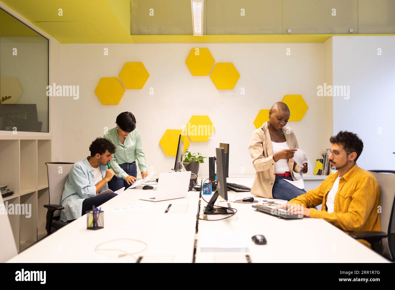Ein Team aus verschiedenen Kollegen, die sich Dokumente anschauen, und Arbeitsmännern, die auf den Monitor schauen, während sie am Tisch sitzen und ethnische Frauen im Büro haben Stockfoto