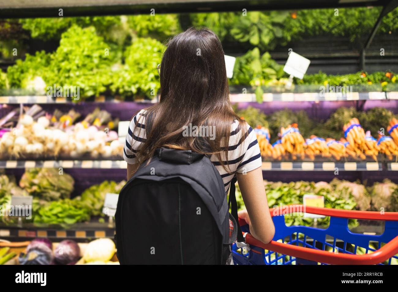Rückansicht einer nicht erkennbaren Kundin mit Rucksack und Warenkorb, die frisches Gemüse in den Regalen im Supermarkt wählt Stockfoto