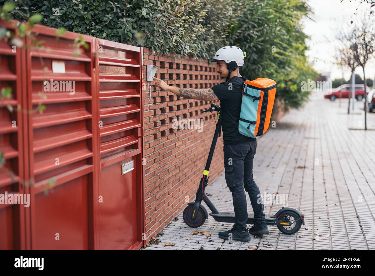 Ganzkörper-Seitenansicht des jungen Lieferers im Helm mit thermischem Rucksack auf elektrischem Tretroller, der die Türklingel des Hauses klingelt Stockfoto