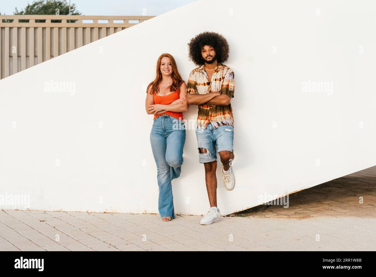 Eine ganze Länge von fröhlichen rothaarigen Frauen und afroamerikanischen Männern mit Afro-Haaren, die die Füße gegen die Wand legen, während sie die Arme kreuzen und in die Kamera schauen Stockfoto