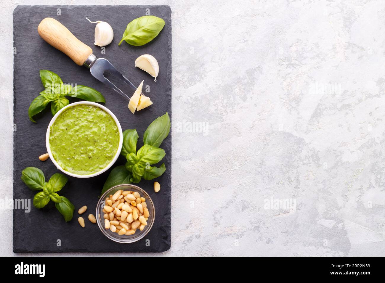 Pesto-Sauce und Zutaten, Käse, Basilikum, Knoblauch, Pinienkerne, Olivenöl auf schwarzem Käseplatte und grauem Steinhintergrund mit Kopierfläche Stockfoto