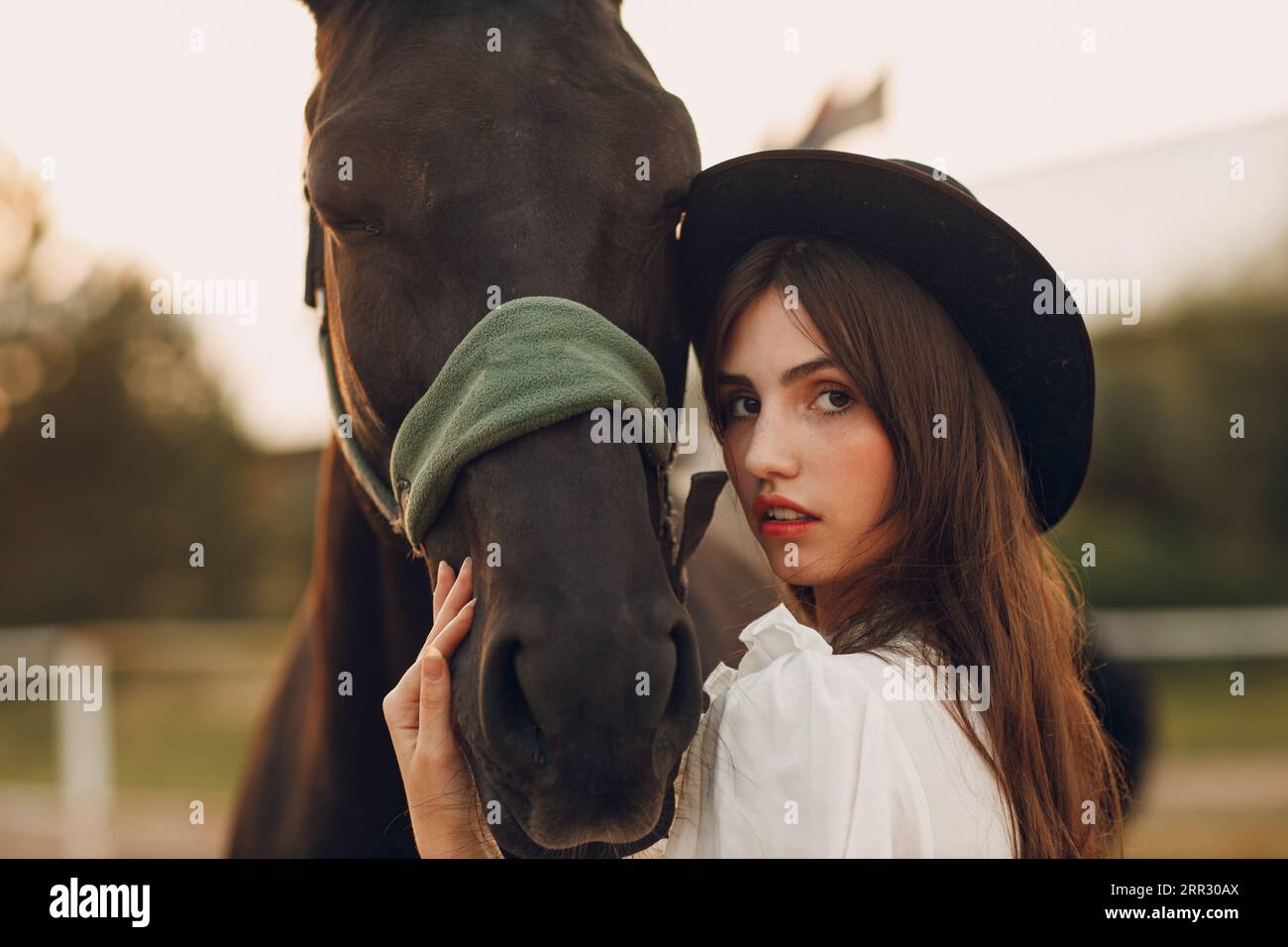 Frau in weißem Kleid und schwarzem Hut mit ihrem Pferd auf der Ranch im Freien Stockfoto