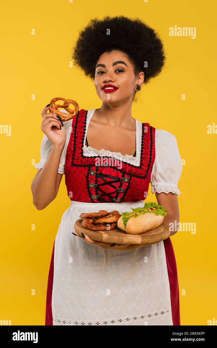 Fröhliche afroamerikanische oktoberfest-Kellnerin mit Brezeln und Hot Dog auf Holztablett auf gelb Stockfoto